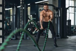 Naked muscular man exercising with battle ropes at gym